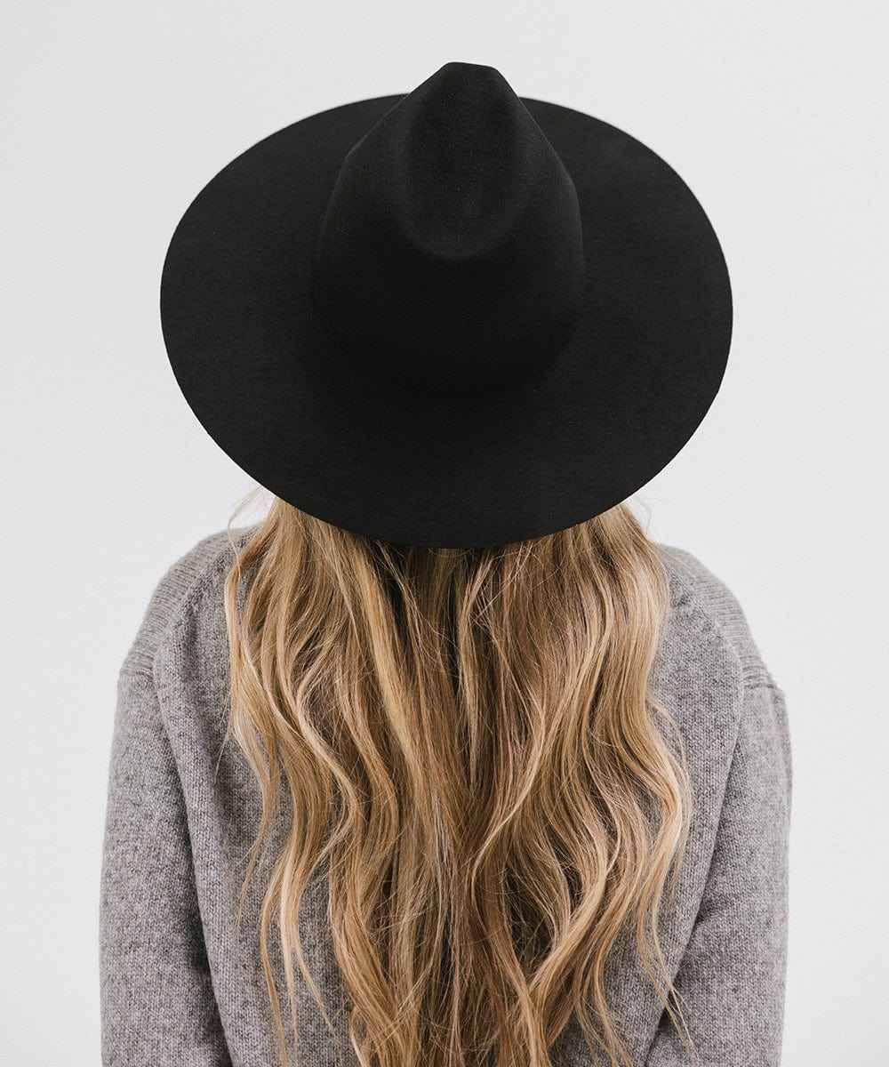 Woman with long blonde hair wearing a black wide brim fedora hat and a grey sweater, facing behind against a plain background #color_black