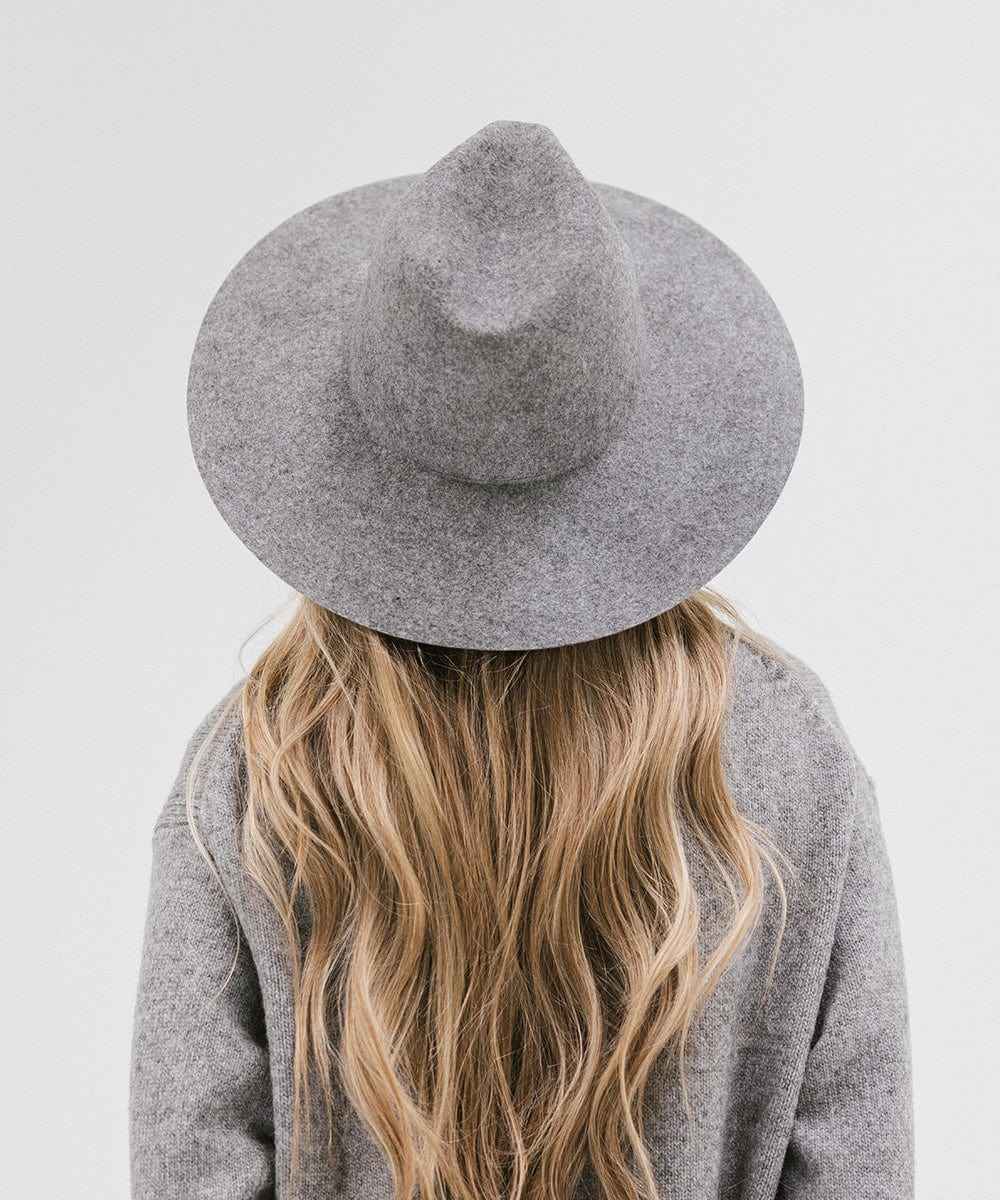 Woman with long blonde hair wearing a grey wide brim fedora hat and a grey sweater, facing behind against a plain background #color_grey