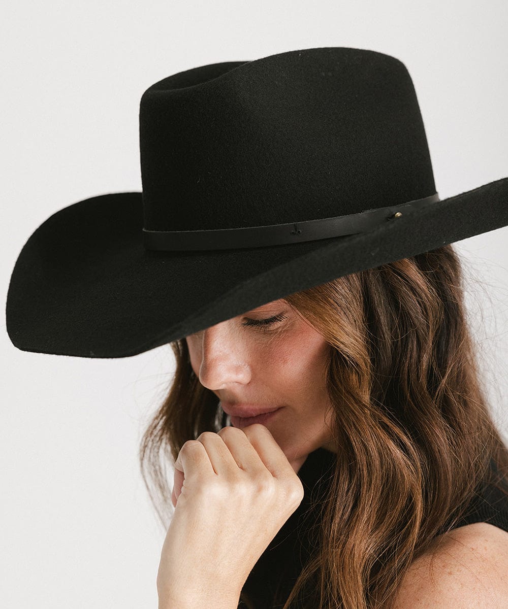 Woman wearing a black cowboy hat with an upturned brim and an attached leather band against a white background #color_black