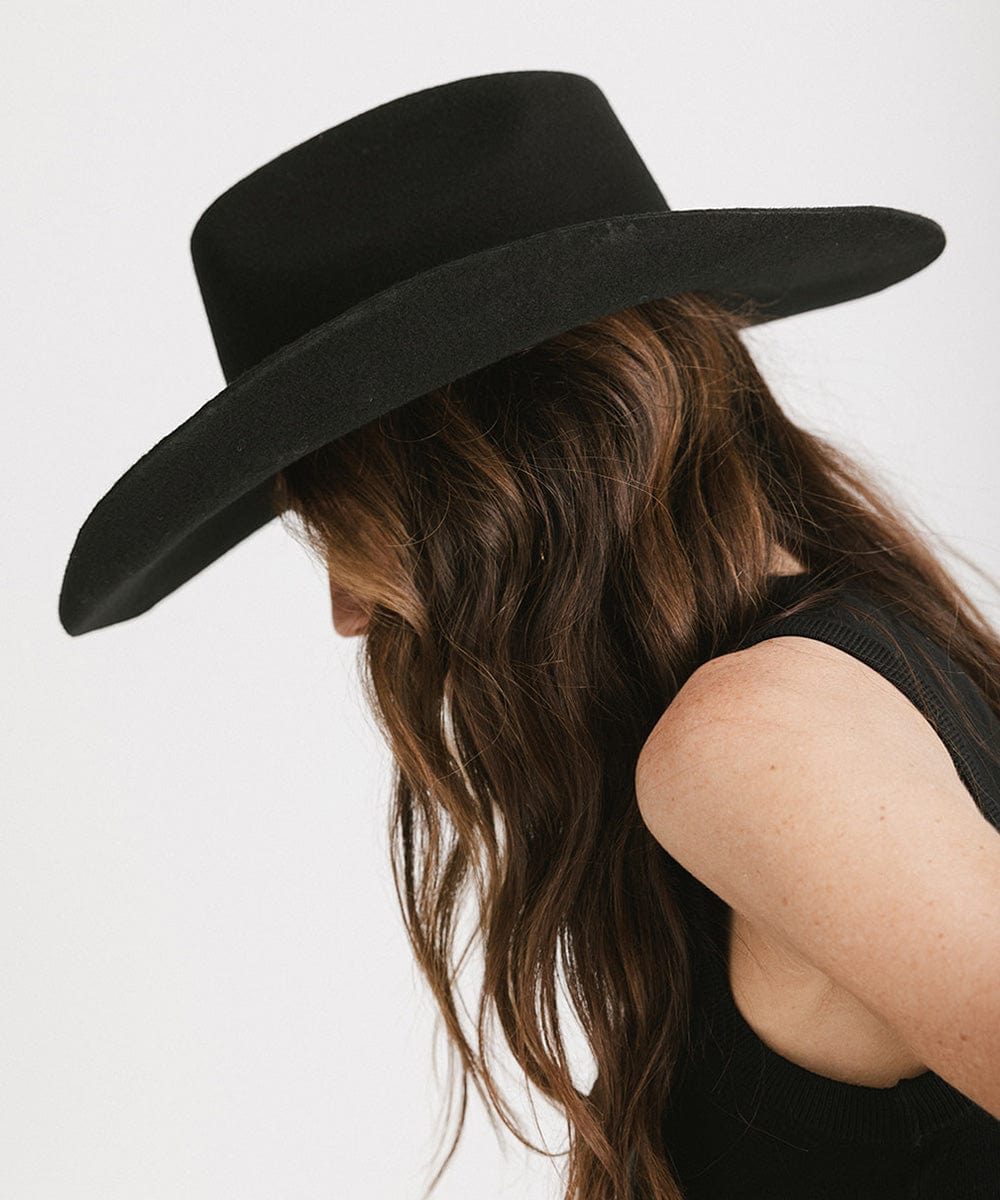 Woman wearing a black cowboy hat with an upturned brim and an attached leather band and a black top, facing sideways against a white background #color_black