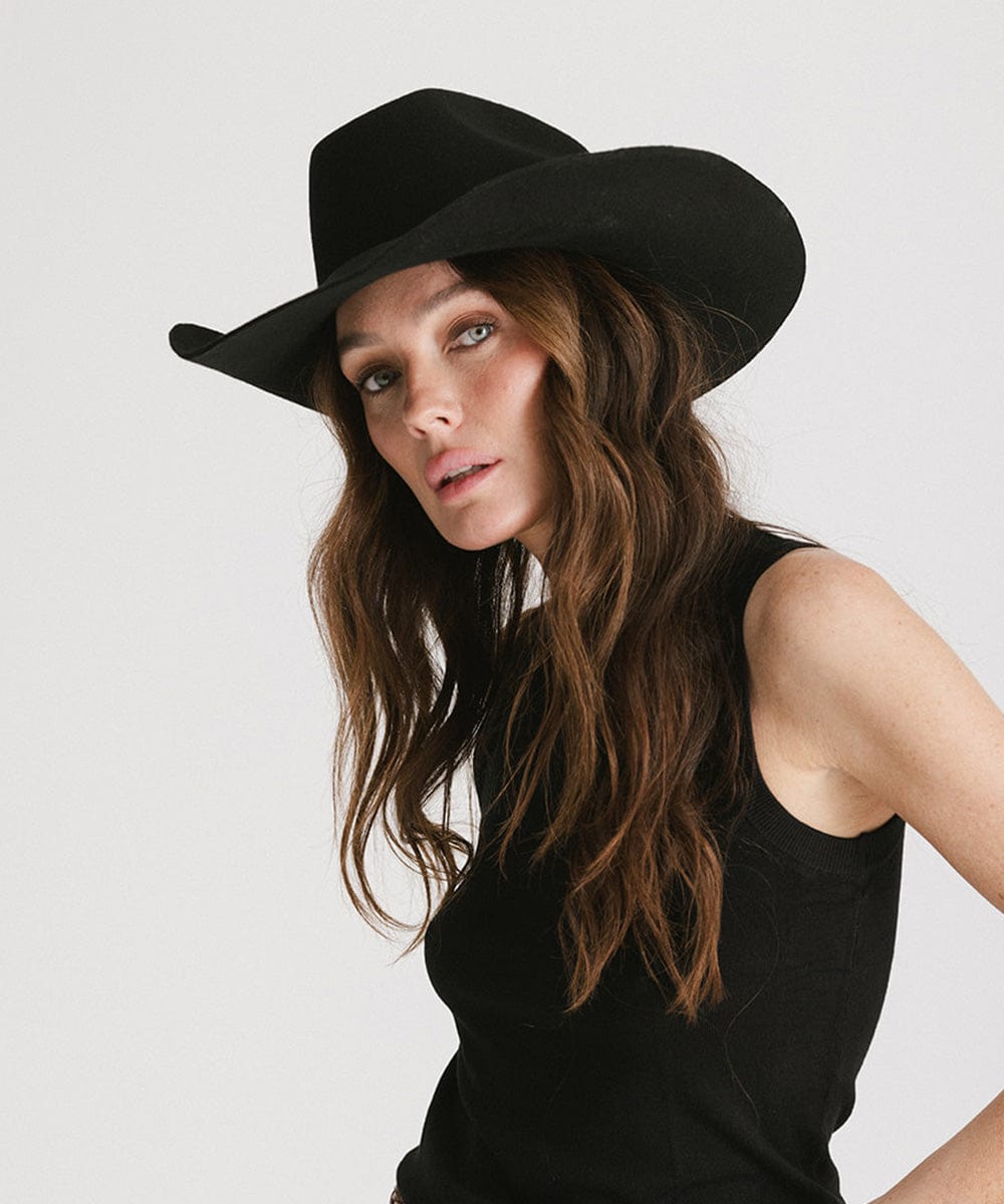 Woman wearing a black cowboy hat with an upturned brim and an attached leather band and a black top against a white background #color_black