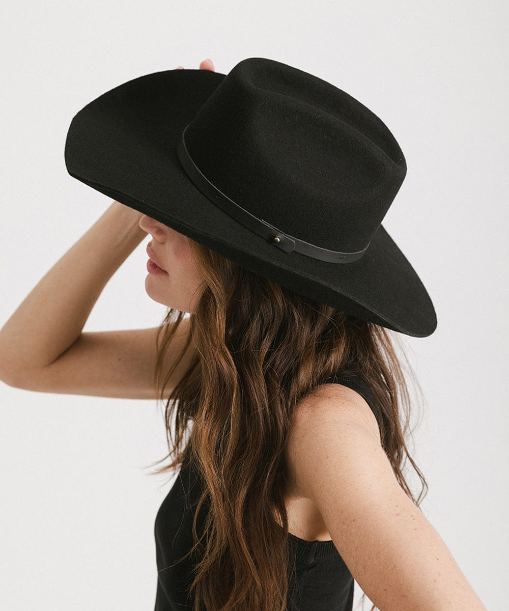 Woman wearing a black cowboy hat with an upturned brim and an attached leather band and a black top, facing sideways against a white background #color_black