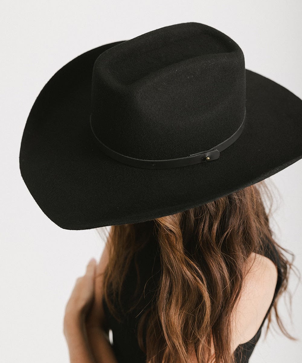 Woman with long hair wearing a black cowboy hat with an upturned brim and an attached leather band and a black top against a white background #color_black