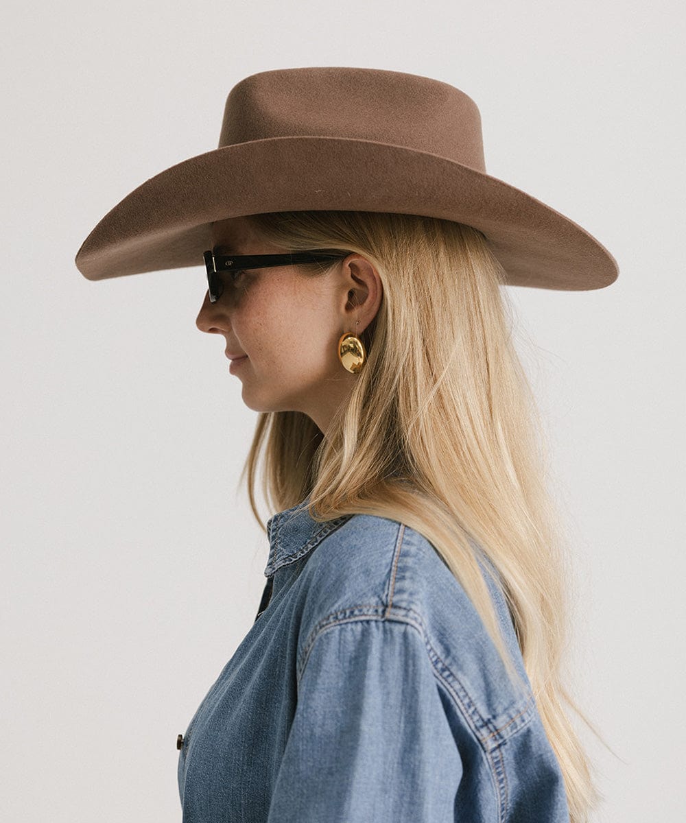 Woman wearing a chocolate colored cowboy hat with an upturned brim and an attached leather band, sunglasses, and a denim shirt, facing sideways against a white background #color_chocolate