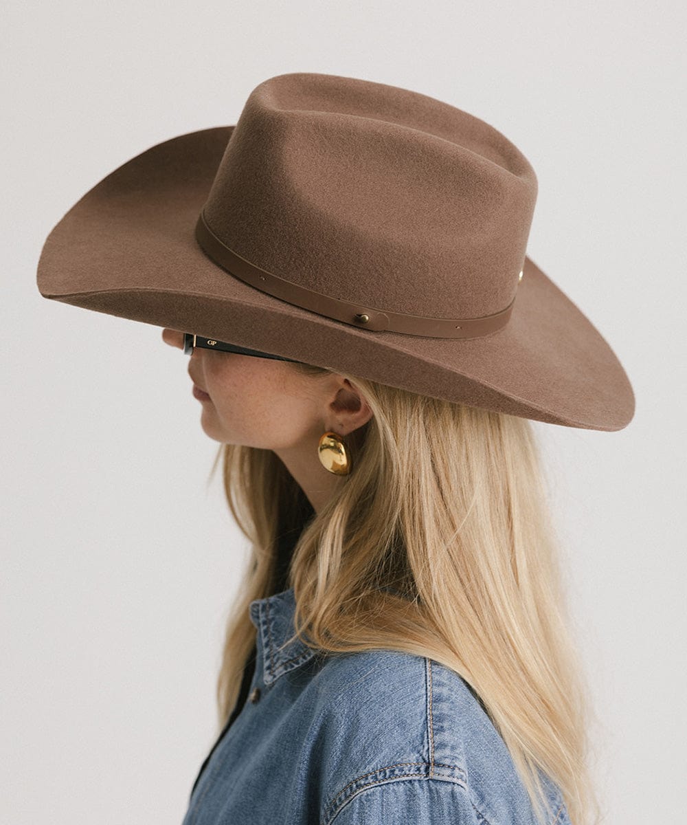Woman wearing a chocolate colored cowboy hat with an upturned brim and an attached leather band, and a denim shirt, facing sideways against a white background #color_chocolate