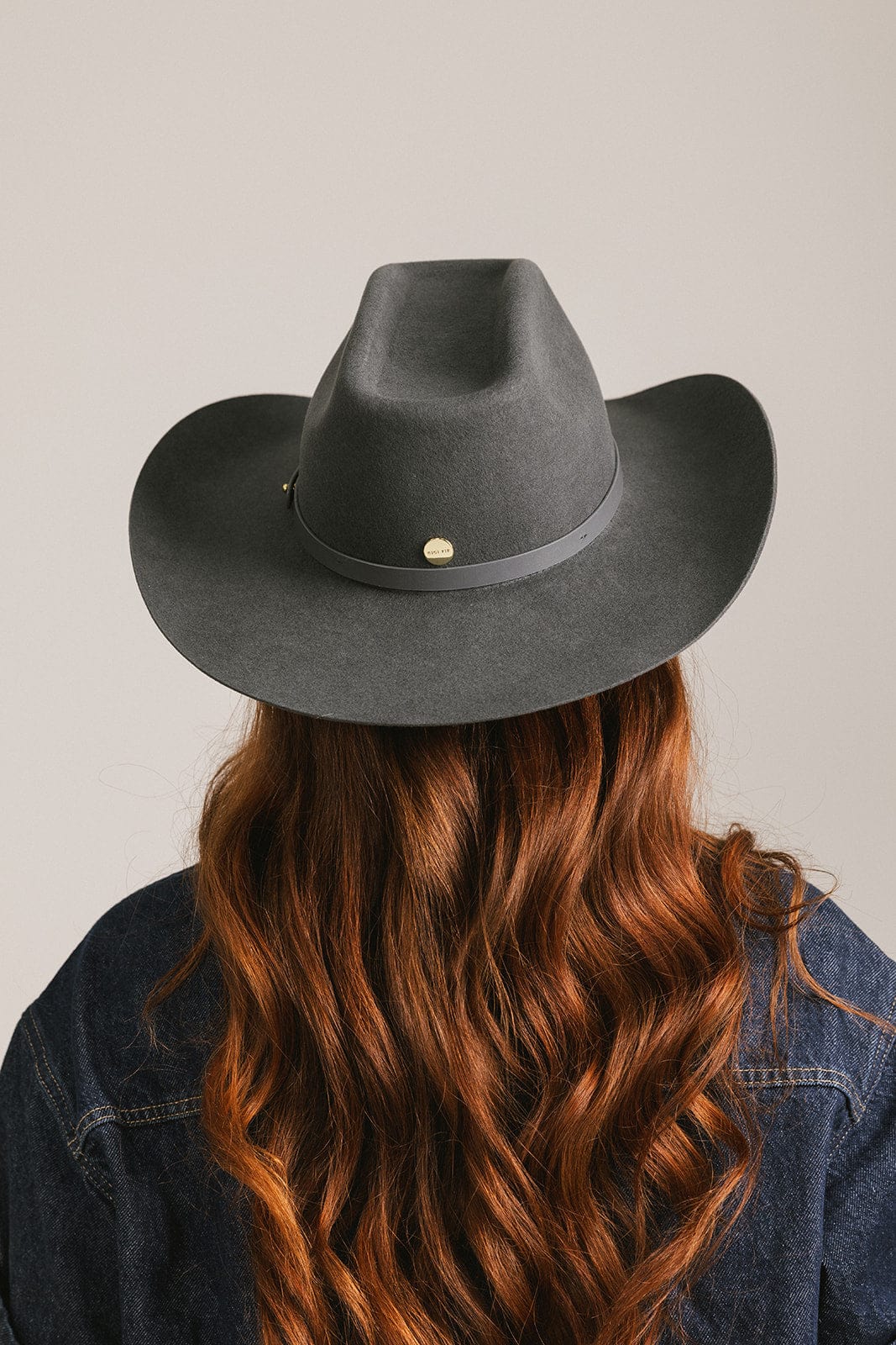 Woman wearing a dark grey cowboy hat with an upturned brim and golden button and an attached leather band, and a dark blue denim shirt, facing behind against a white background #color_dark grey