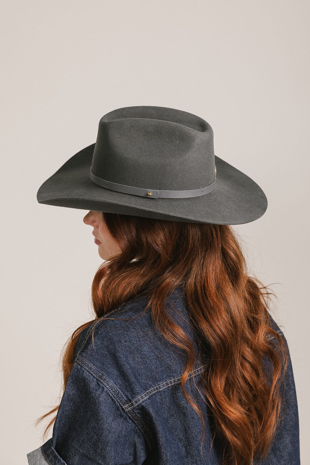 Woman wearing a dark grey cowboy hat with an upturned brim and an attached leather band and a dark blue denim shirt, facing sideways against a white background #color_dark grey