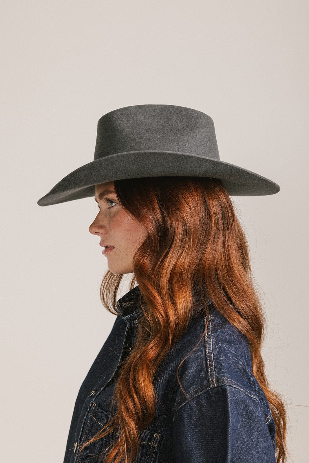 Woman wearing a dark grey cowboy hat with an upturned brim and an attached leather band and a dark blue denim shirt, facing sideways against a white background #color_dark grey