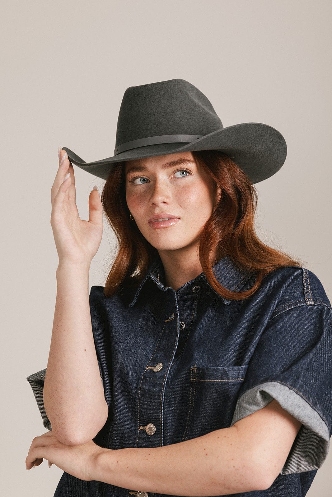 Woman wearing a dark grey cowboy hat with an upturned brim and an attached leather band and a dark blue denim shirt, looking sideways against a white background #color_dark grey