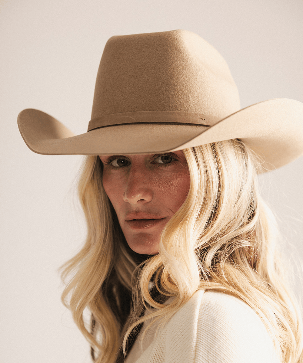 A close-up portrait of a woman wearing a tan felt cowboy hat, looking directly at the camera against a plain light background #color_tan
