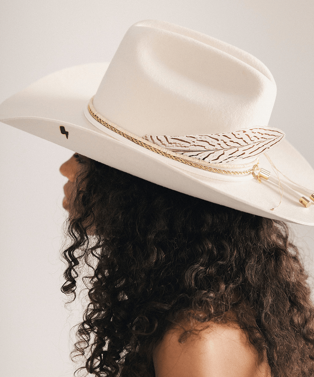 Woman wearing a white cowboy hat with a lightning brand, decorative band, and feather on a plain background #color_off white