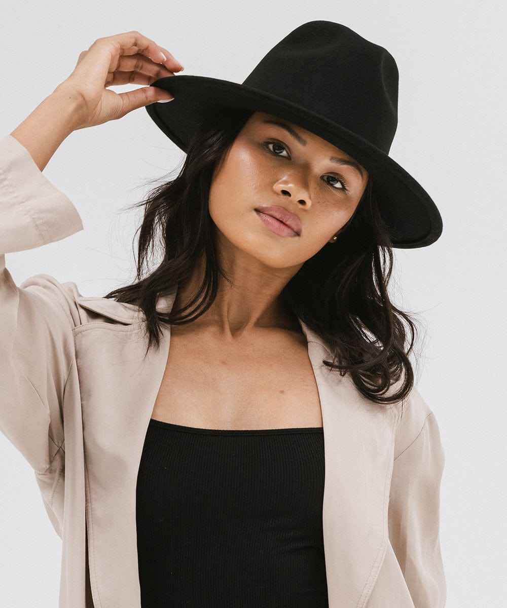 Woman wearing a black fedora hat and a beige coat with a black top against a plain background #color_black