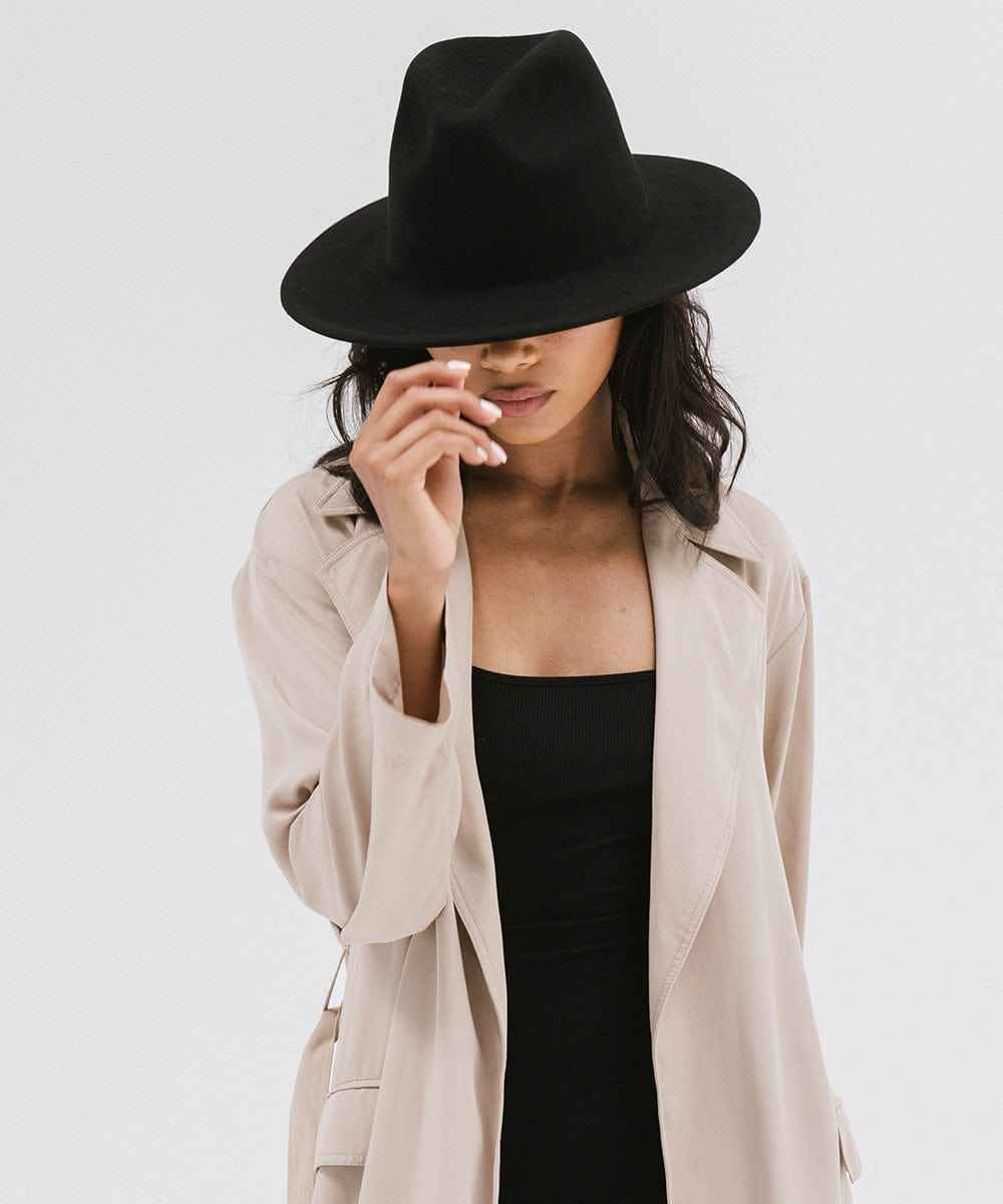 Woman wearing a black fedora hat and a beige coat with a black top against a plain background #color_black