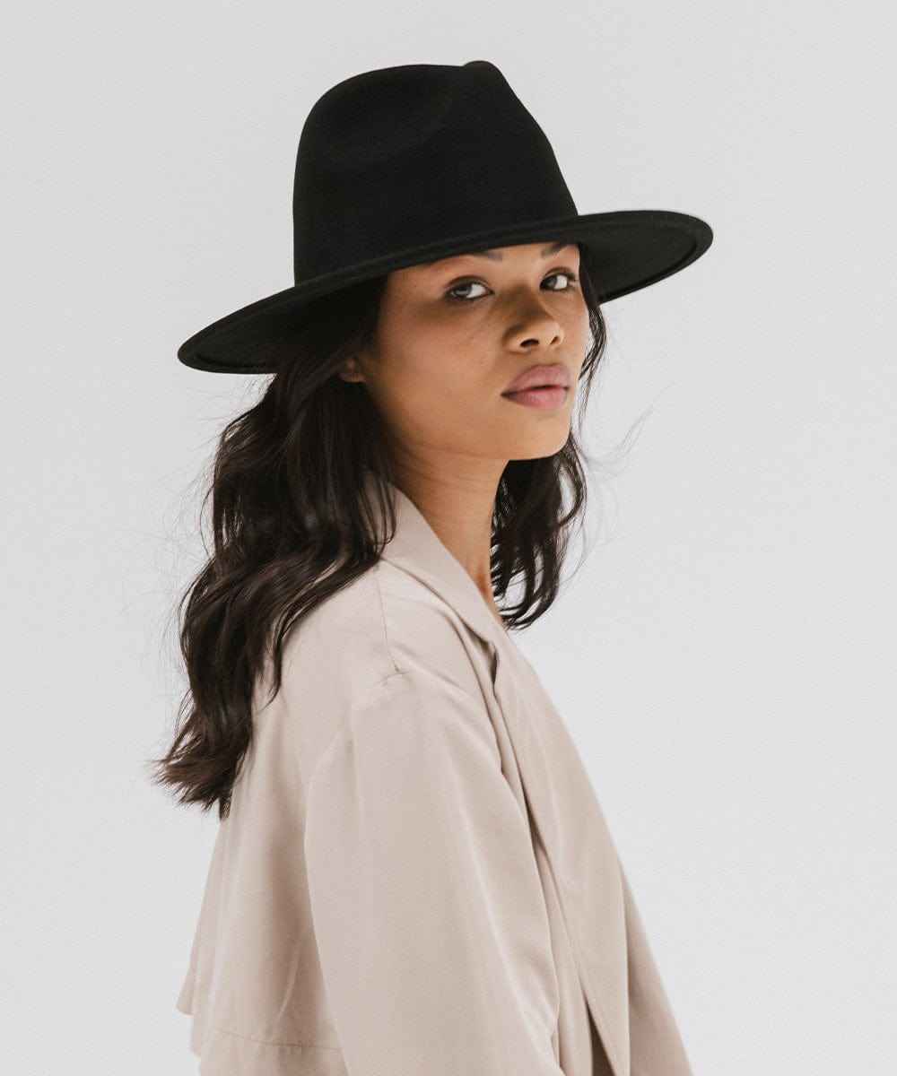 Woman wearing a black fedora hat and a beige coat against a plain background #color_black