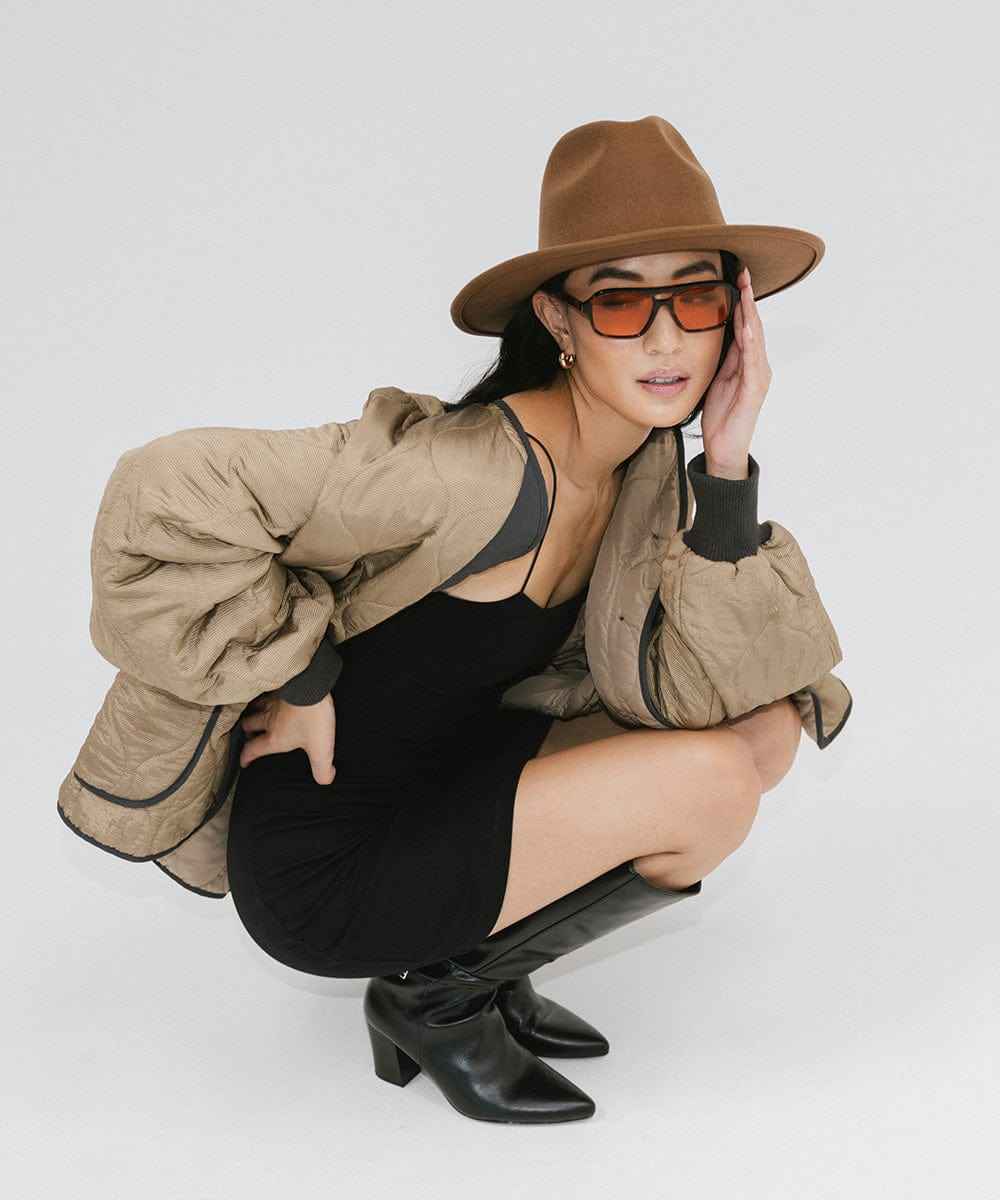 Woman wearing a brown fedora hat, sunglasses, a black top and a beige jacket, and black boots against a plain background #color_brown