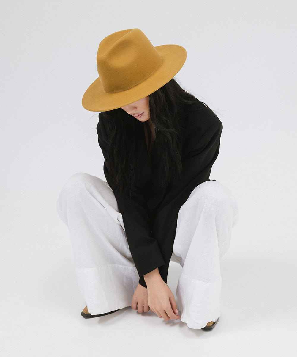 Woman wearing a mustard colored fedora hat, a black coat and white pants, sitting against a plain background #color_mustard