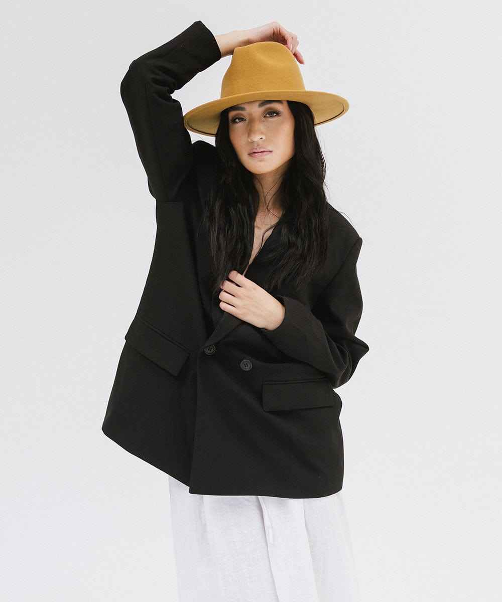 Woman wearing a mustard colored fedora hat and a black coat and white pants against a plain background #color_mustard