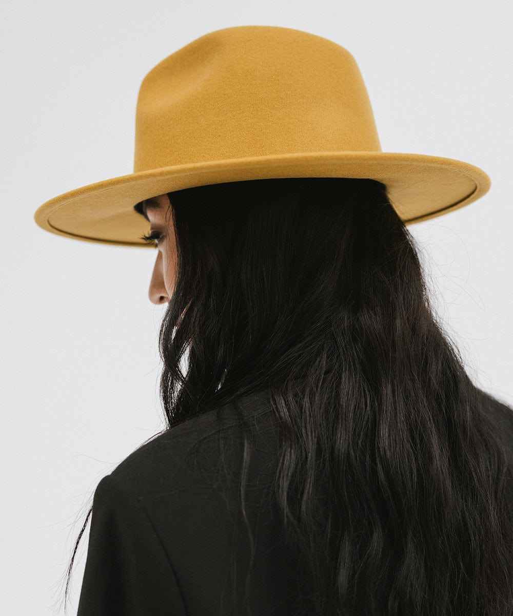 Woman wearing a mustard colored fedora hat and a black coat against a plain background #color_mustard