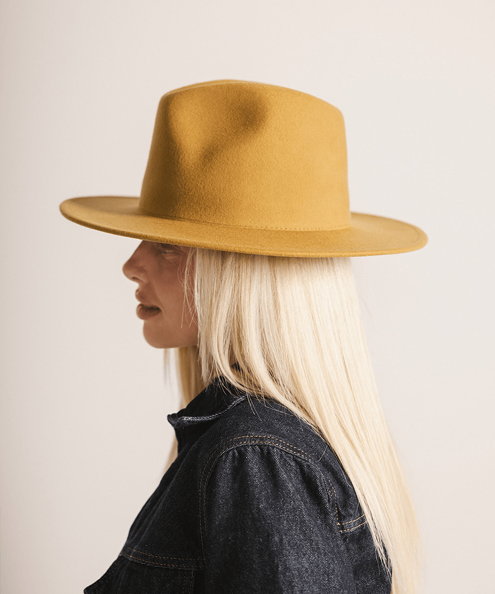 Woman wearing a mustard colored fedora hat and a black coat against a plain background #color_mustard