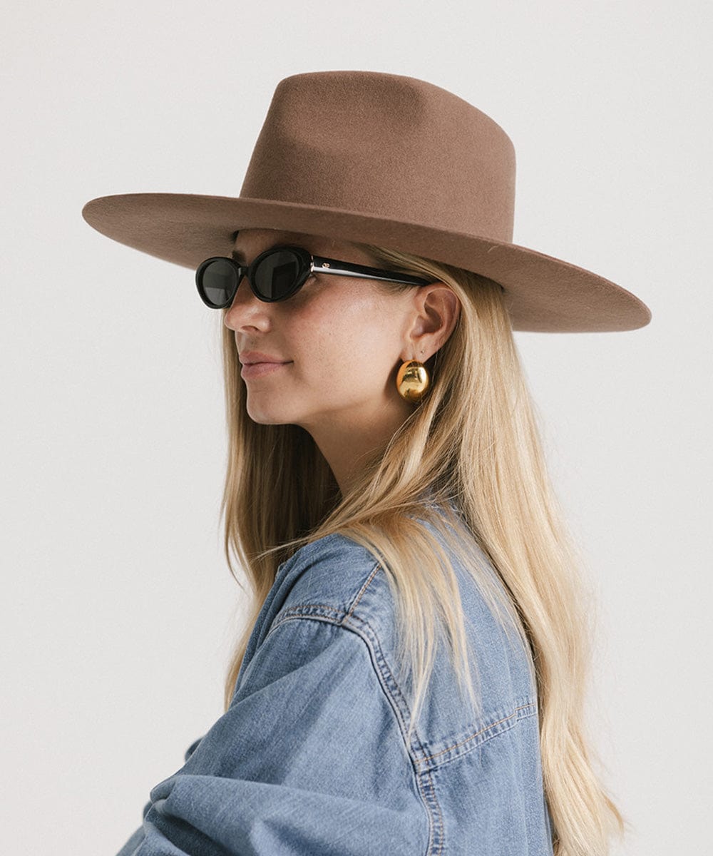 Woman wearing a chocolate colored fedora hat, sunglasses and a denim shirt, looking sideways against a plain background #color_chocolate