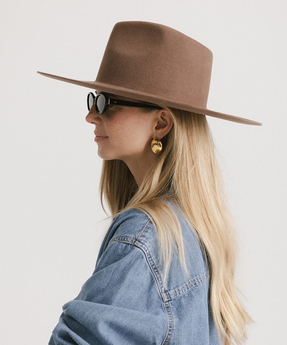 Woman wearing a chocolate colored fedora hat, sunglasses and a denim shirt, facing sideways against a plain background #color_chocolate