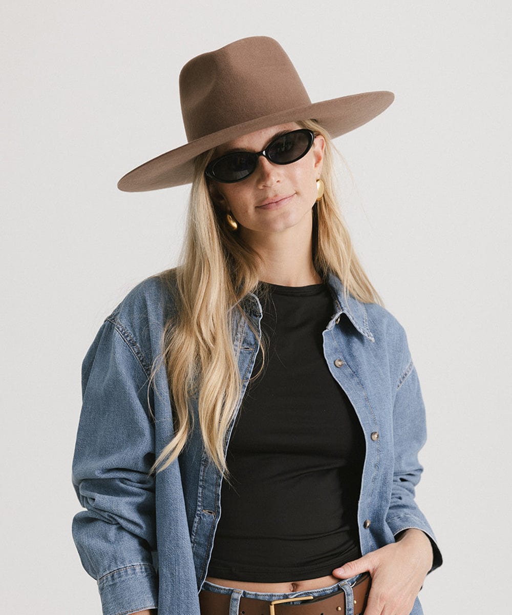 Woman wearing a chocolate colored fedora hat, sunglasses and a black t-shirt with a denim shirt against a plain background #color_chocolate