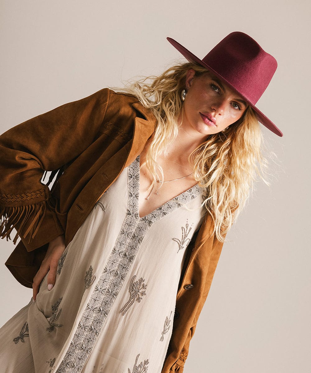 Woman wearing a brown jacket, white dress with black patterns, and a merlot fedora hat against a plain background #color_merlot