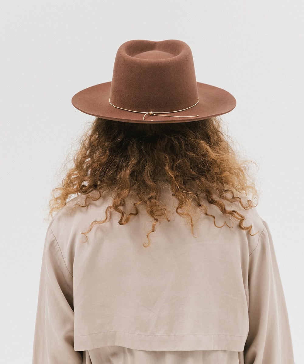 Woman wearing a dark oak colored fedora hat with a hat band and a beige jacket, facing behind against a white background #color_dark oak