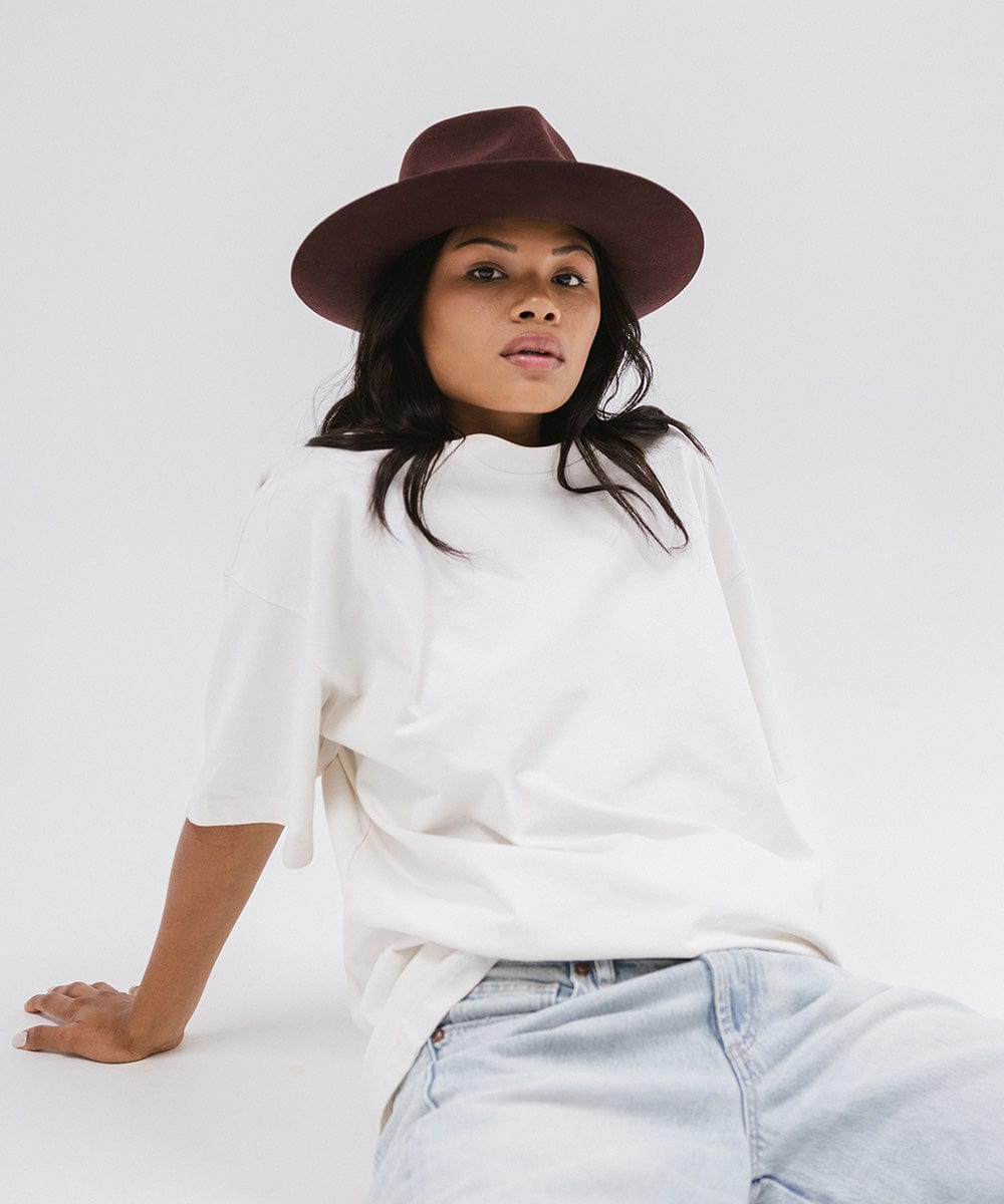 Woman wearing a dark cherry colored fedora hat and a white shirt, sitting against a white background #color_dark cherry