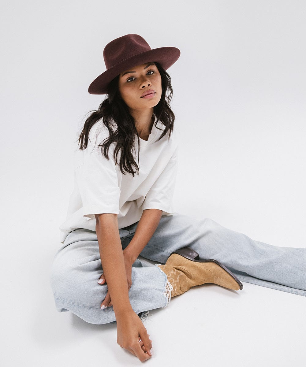 Woman wearing a dark cherry colored fedora hat, a white shirt, blue jeans and brown boots, sitting against a white background #color_dark cherry