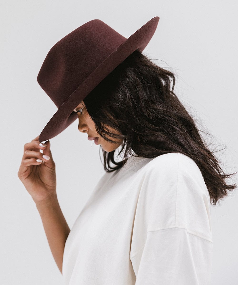 Woman wearing a dark cherry colored fedora hat and a white shirt, posing sideways against a white background #color_dark cherry