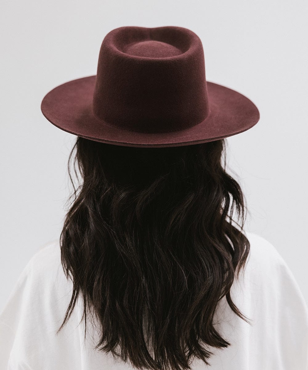 Woman wearing a dark cherry colored fedora hat and a white shirt, facing behind against a white background #color_dark cherry