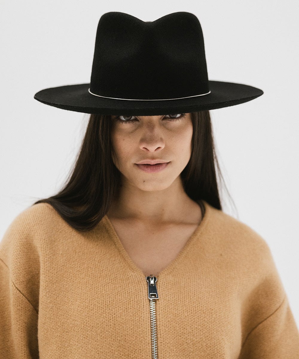 Woman wearing a black fedora hat with a band and a brown jacket against a white background #color_black