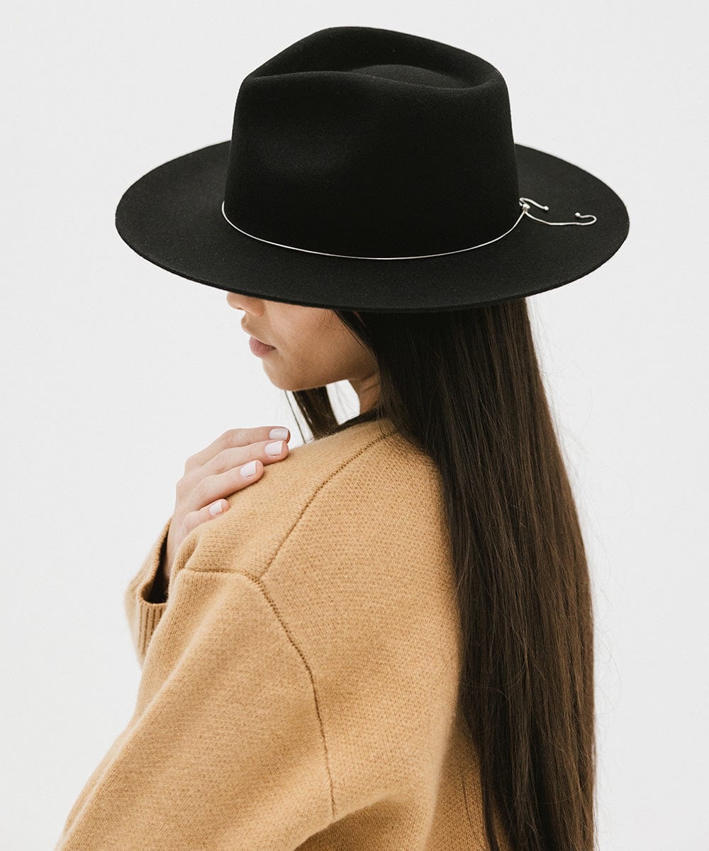 Woman wearing a black fedora hat with a band and a brown jacket, facing sideways against a white background #color_black