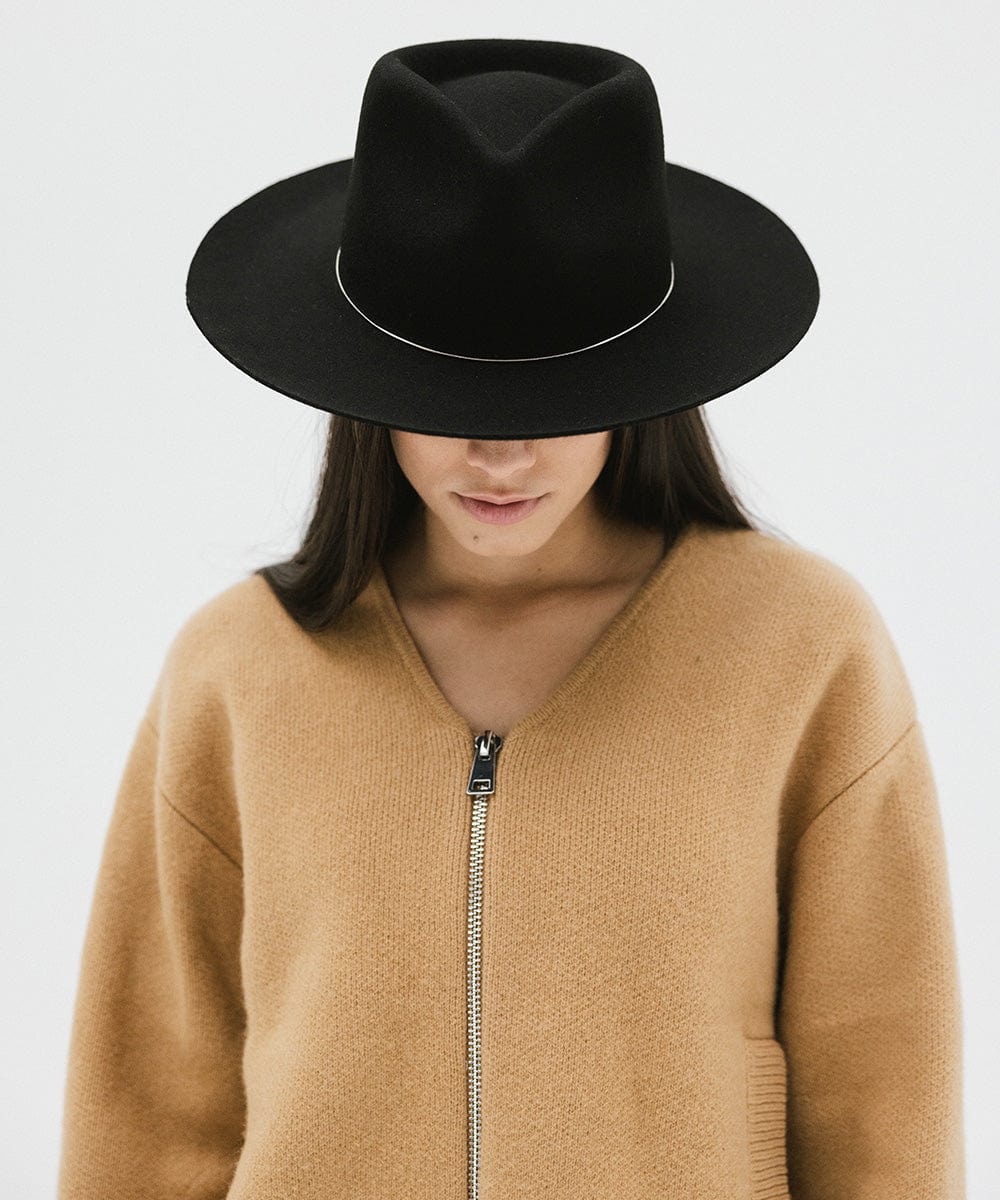 Woman wearing a black fedora hat with a band and a brown jacket, looking down against a white background #color_black