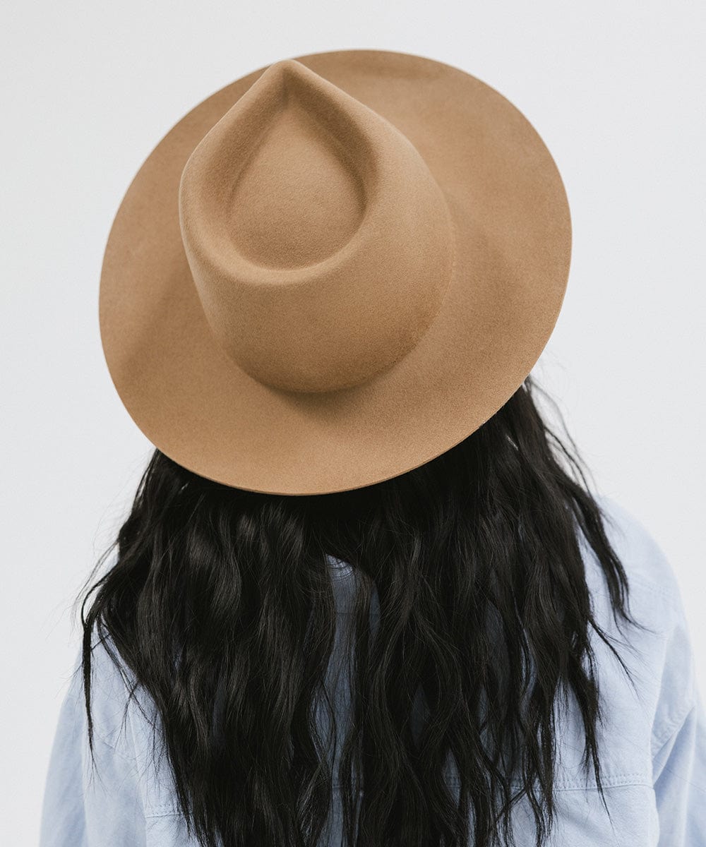 Woman wearing a brown fedora hat and a light blue shirt, facing behind against a white background #color_brown