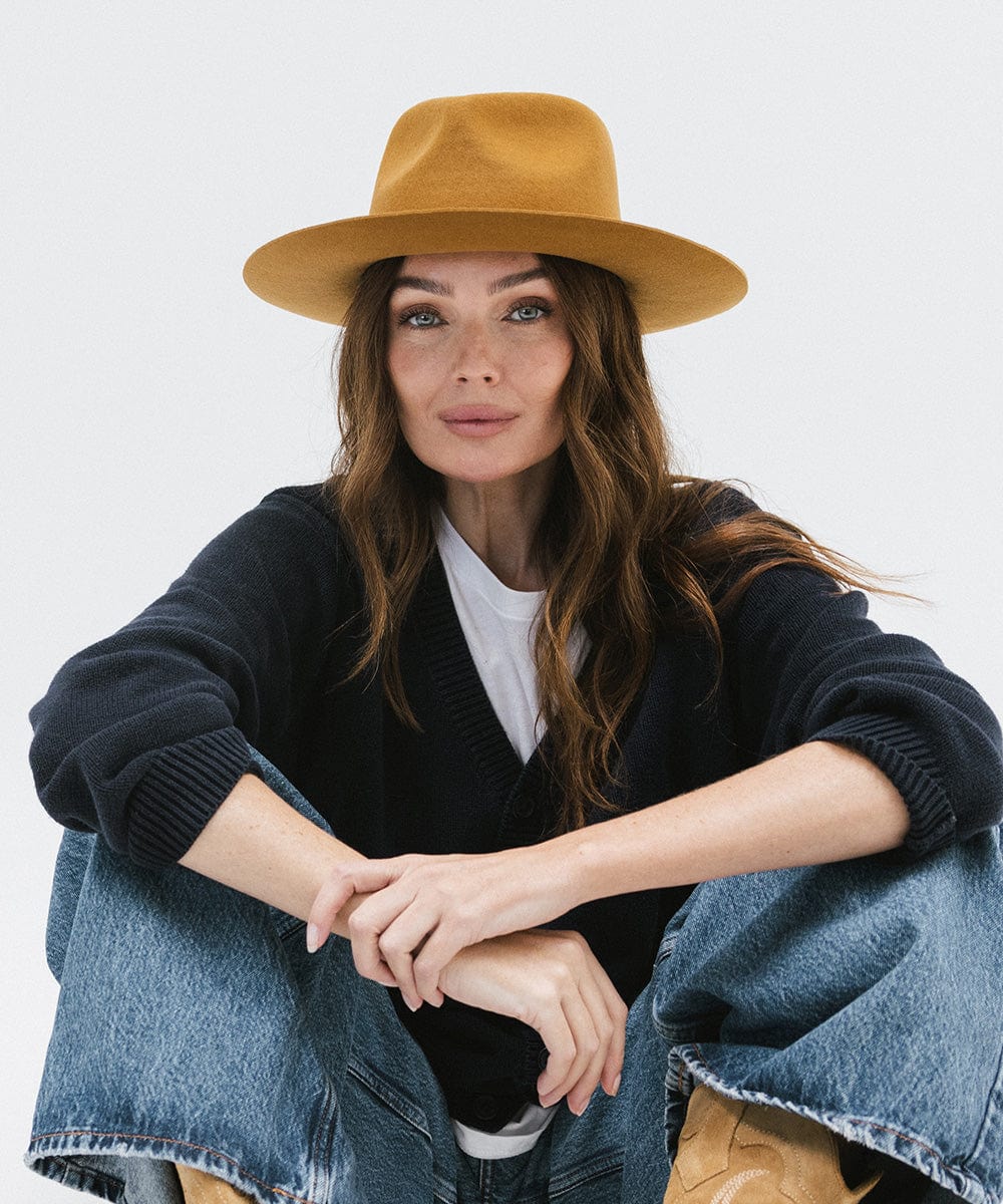 Woman wearing a cinnamon colored fedora hat, a dark blue shirt and blue jeans, sitting against a white background #color_cinnamon
