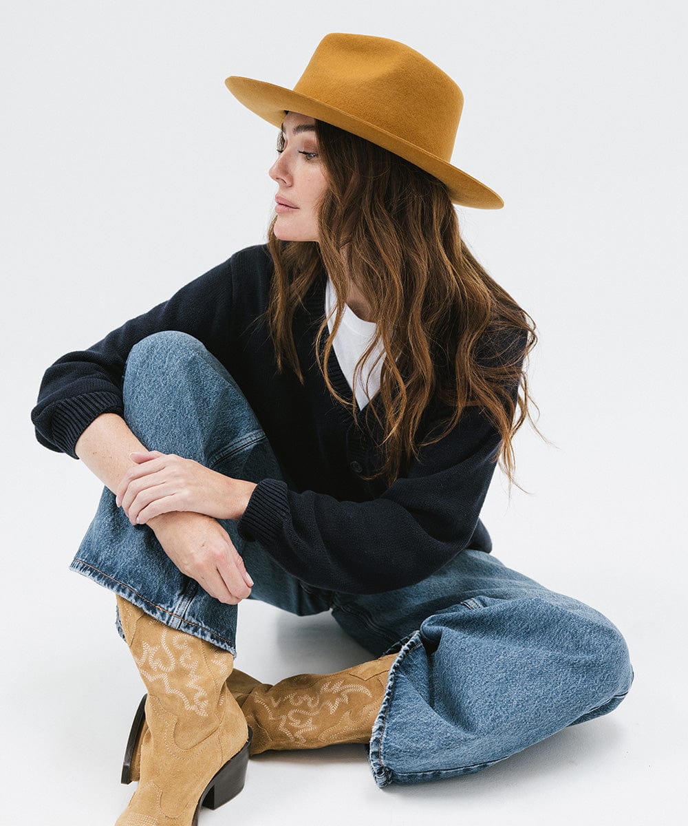 Woman wearing a cinnamon colored fedora hat, a dark blue shirt, blue jeans, and brown boots, sitting against a white background #color_cinnamon