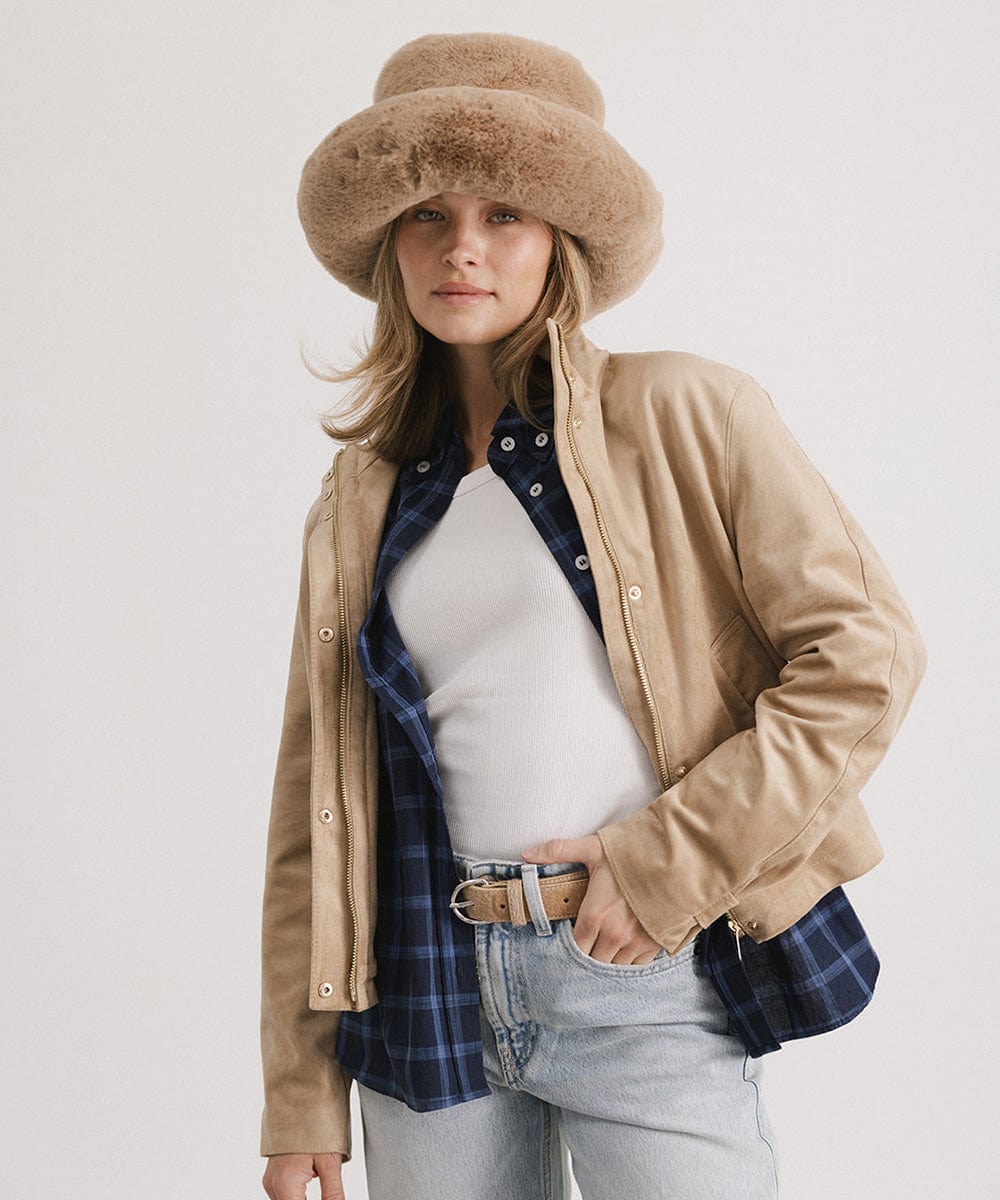 Woman wearing a tan faux fur bucket hat, a tan jacket with a blue shirt and white top, and light blue jeans, against a plain background #color_tan