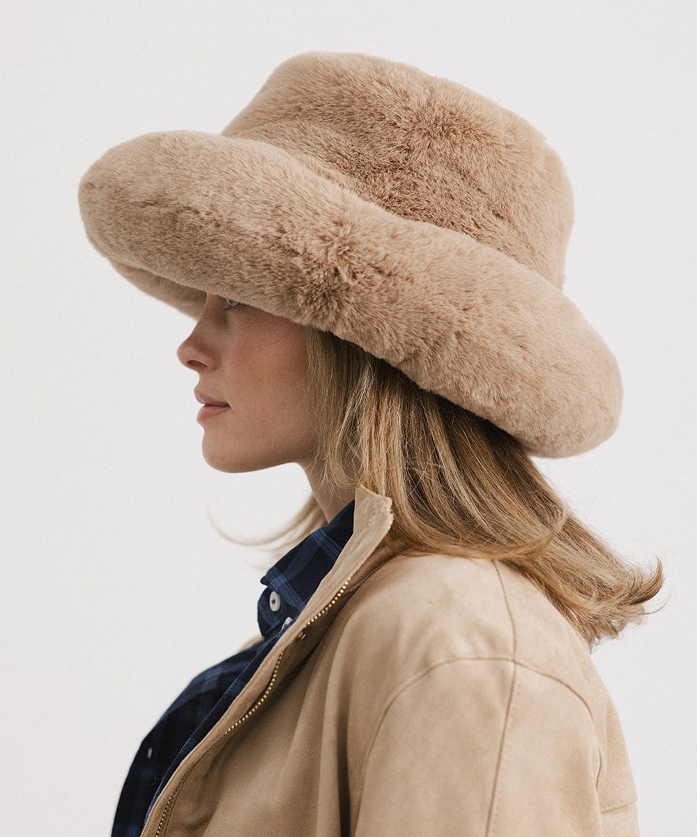 Woman wearing a tan faux fur bucket hat and a tan jacket, facing sideways  against a plain background #color_tan