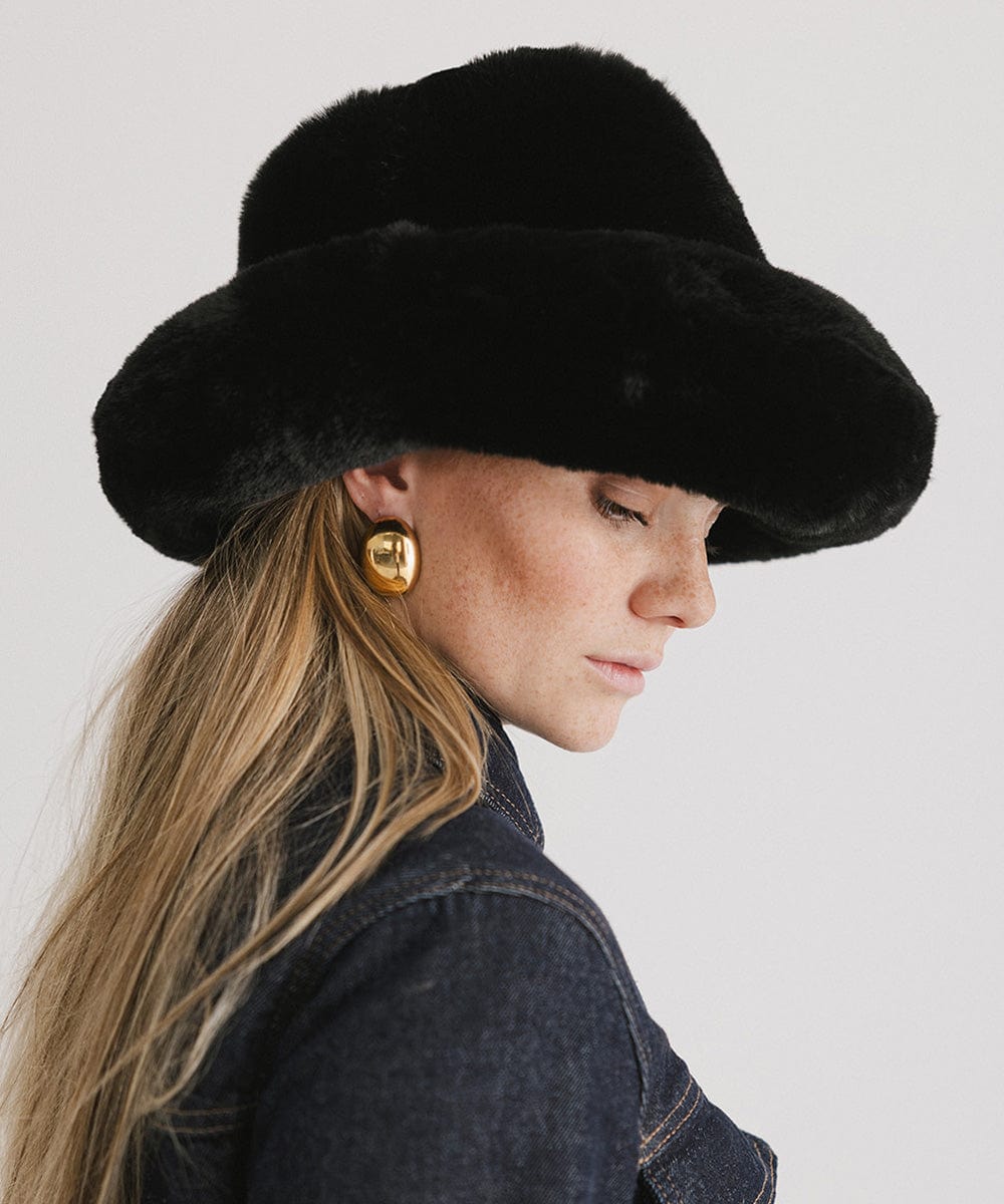 Woman wearing a black faux fur bucket hat and a denim shirt, facing sideways against a plain background #color_black