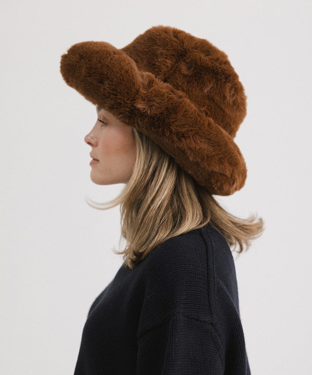 Woman wearing a brown faux fur bucket hat and a dark blue sweater, facing sideways against a plain background #color_brown