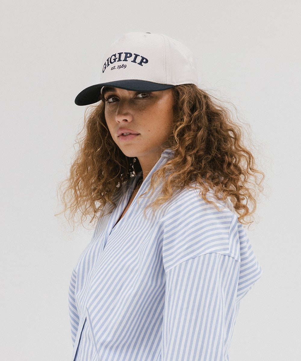 Woman wearing a cream and navy  trucker hat with 'GIGI PIP' embroidered in navy on the front and a light colored shirt, posing sideways  against a plain background #color_cream - navy