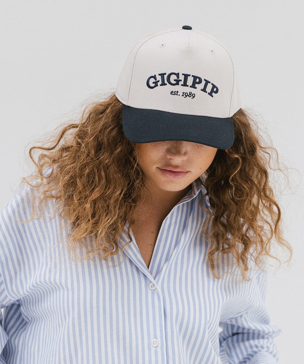 Woman wearing a cream and navy  trucker hat with 'GIGI PIP' embroidered in navy on the front and a light colored shirt against a plain background #color_cream - navy