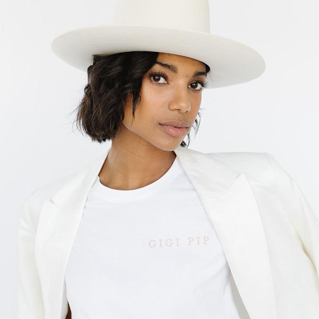 Woman wearing a white t-shirt with 'GIGI PIP' text, a white blazer and a cream hat against a white background #color_white