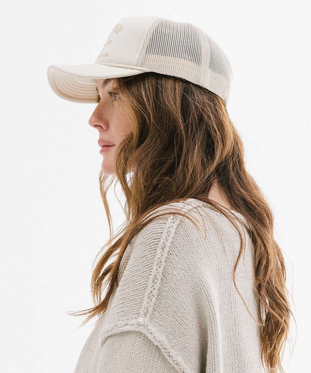 Woman wearing a cream cap with golden 'GIGIRODEO' text and logo, and a grey sweater, facing sideways  against a plain background #color_cream