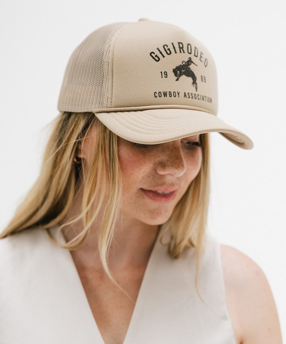 Woman wearing a tan cap with golden 'GIGIRODEO' text and logo, and a white top against a plain background  #color_tan