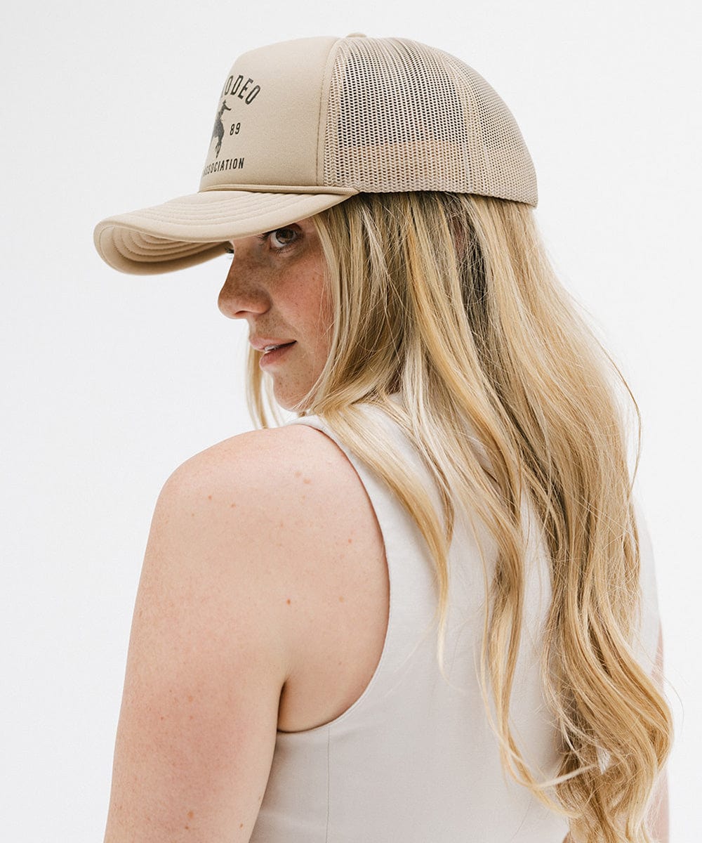 Woman wearing a tan cap with golden 'GIGIRODEO' text and logo, and a white top, facing sideways  against a plain background  #color_tan
