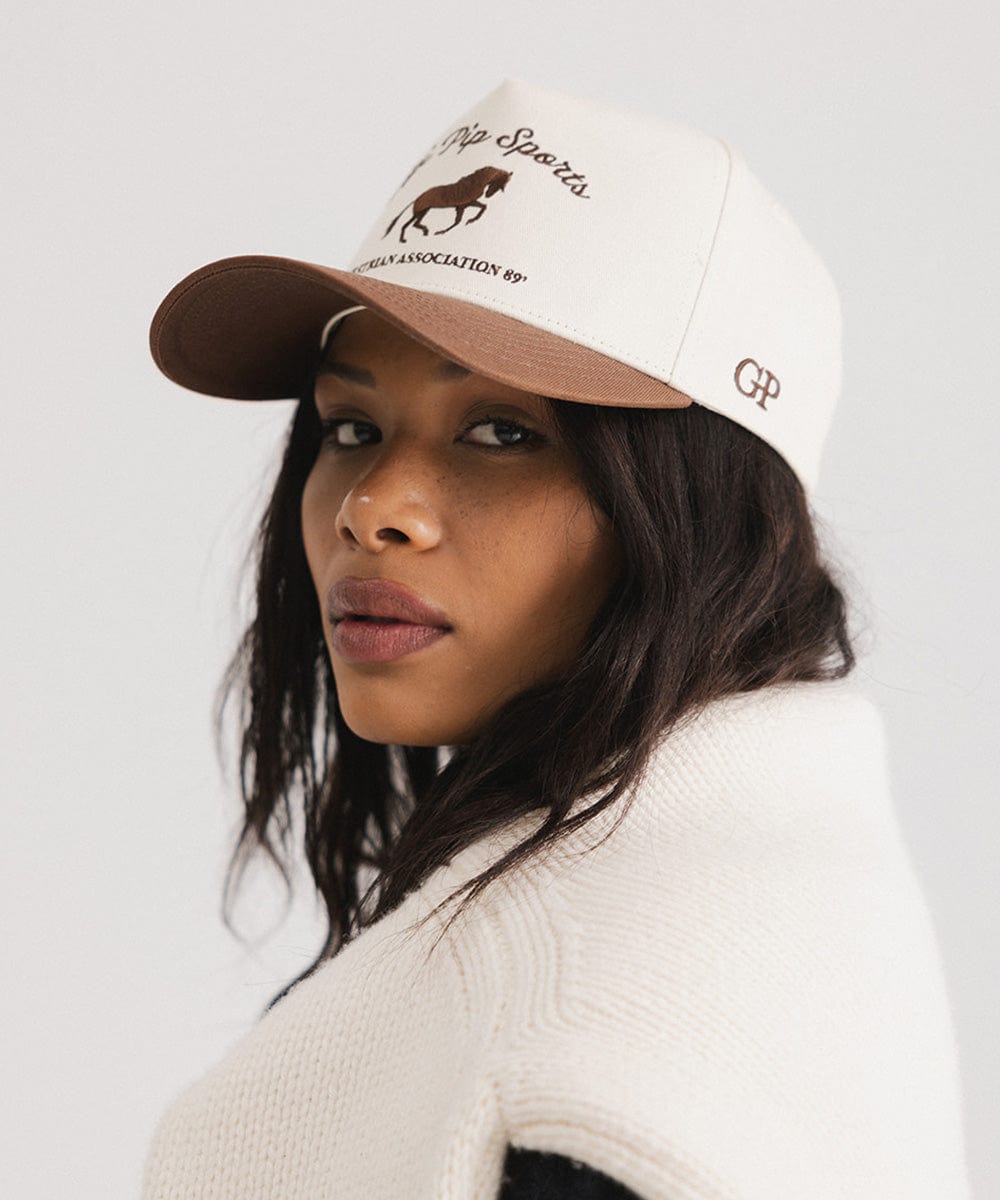 Woman wearing a cream and chocolate brown Trucker hat with 'Gigi Pip Sports' and horse logo and a denim jacket, posing sideways  against a plain background #color_cream - chocolate brown