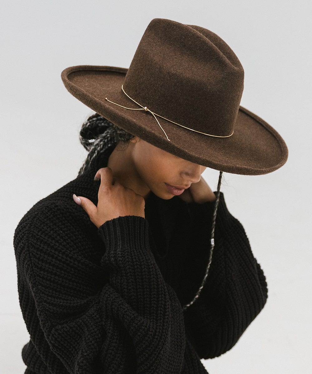 Woman wearing a brown hat with a gold chain bolo hat band and a black sweater, posing sideways against a plain background  #color_gold